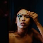 Unemotional ethnic female with bare shoulders reflecting in mirror while removing dark body art makeup from face by using cleansing wipe in dark studio