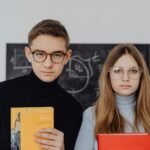 Two students with eyeglasses holding books, standing in front of a chalkboard in a classroom setting.