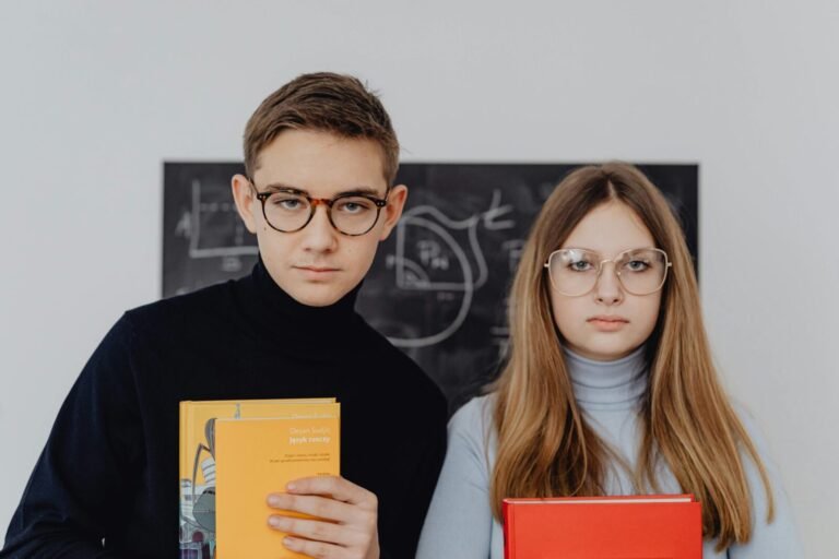 Two students with eyeglasses holding books, standing in front of a chalkboard in a classroom setting.